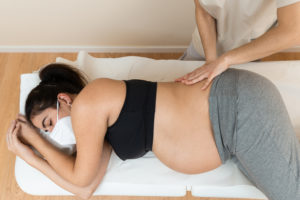 A pregnant woman wearing face mask lying on her side on a massage table while an acupuncturist massages